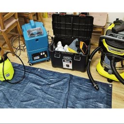 Joanna Kędzierska Fast Cleaning - Profesjonalny sprzęt do sprzątania: odkurzacz Karcher, szorowarka, spryskiwacz i pojemnik z chemią na niebieskiej plandece, gotowe do użycia w pomieszczeniu.