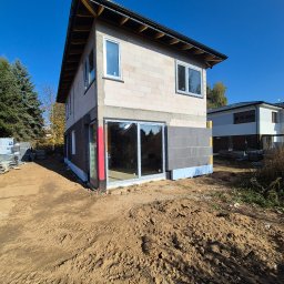 EKIEROWNIK SPÓŁKA Z OGRANICZONĄ ODPOWIEDZIALNOŚCIĄ - Dom w trakcie budowy z pustaków, częściowo ocieplony, z widocznymi oknami i drzwiami balkonowymi. Na pierwszym planie teren budowy z ziemią i śladami opon.