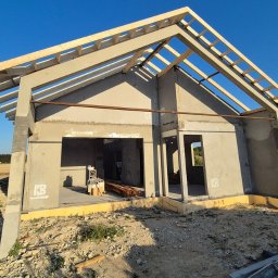 EKIEROWNIK SPÓŁKA Z OGRANICZONĄ ODPOWIEDZIALNOŚCIĄ - Surowy stan domu z betonu komórkowego z drewnianą konstrukcją dachu w trakcie budowy, widok z zewnątrz na tle pola i błękitnego nieba.