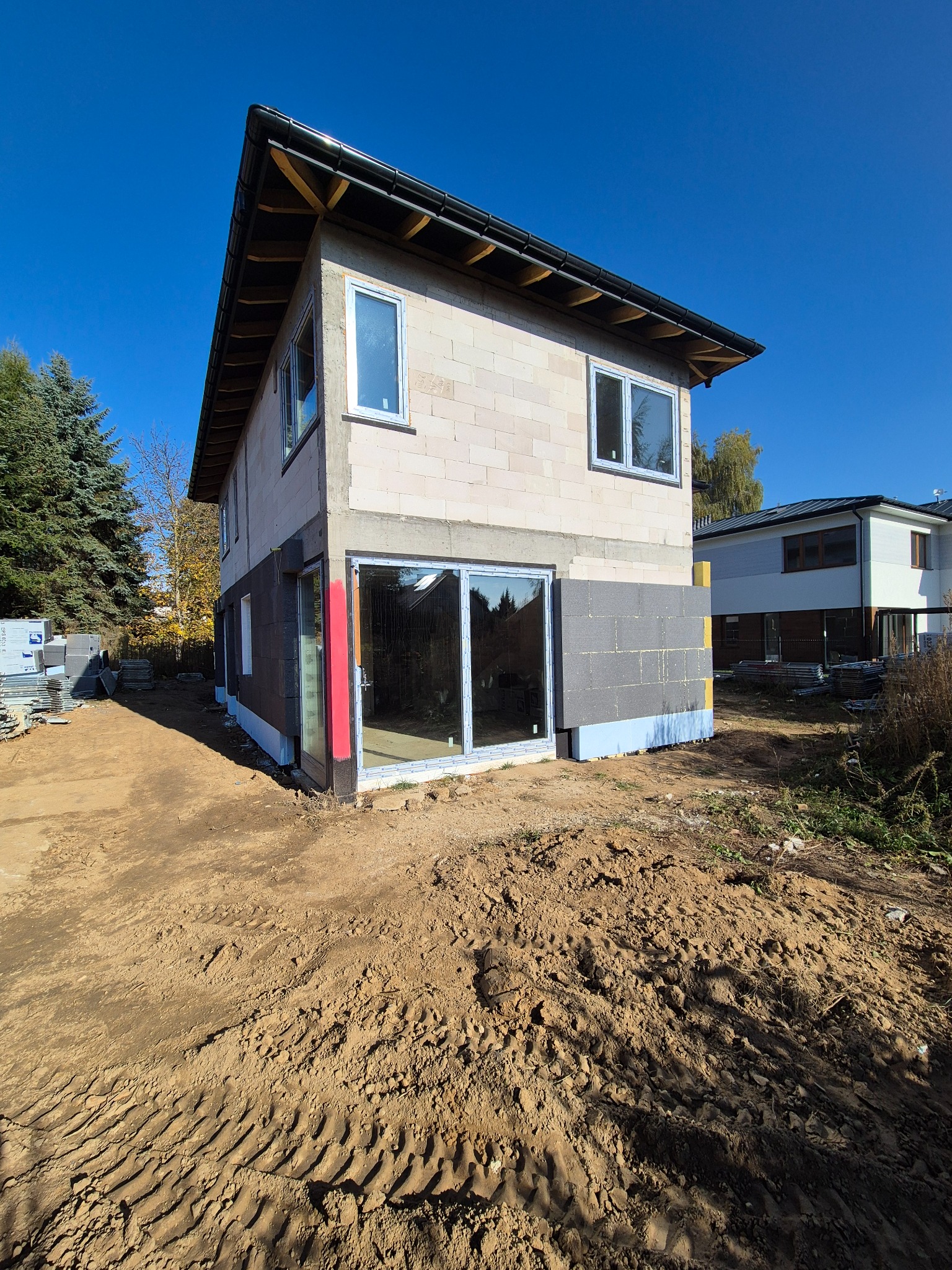 Dom w trakcie budowy z pustaków, częściowo ocieplony, z widocznymi oknami i drzwiami balkonowymi. Na pierwszym planie teren budowy z ziemią i śladami opon.