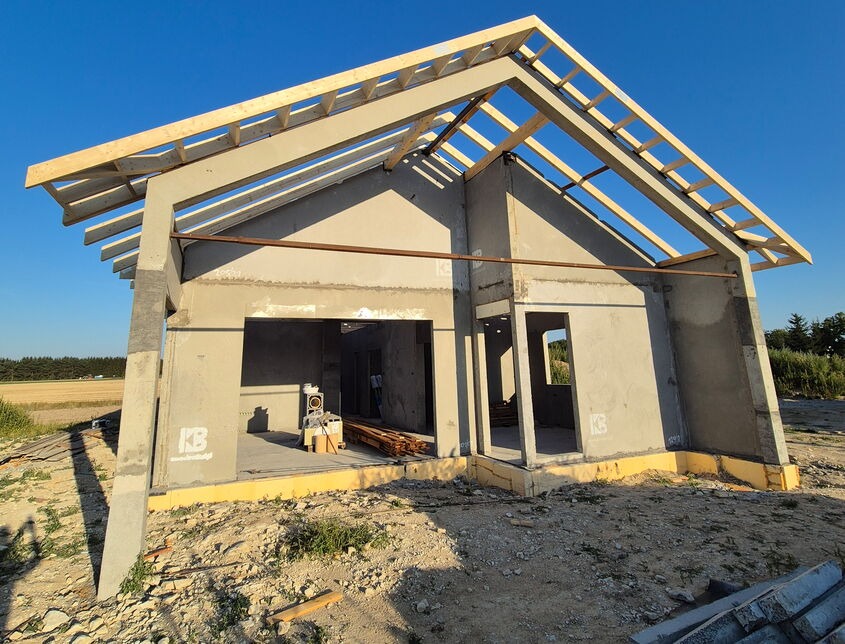 Surowy stan domu z betonu komórkowego z drewnianą konstrukcją dachu w trakcie budowy, widok z zewnątrz na tle pola i błękitnego nieba.