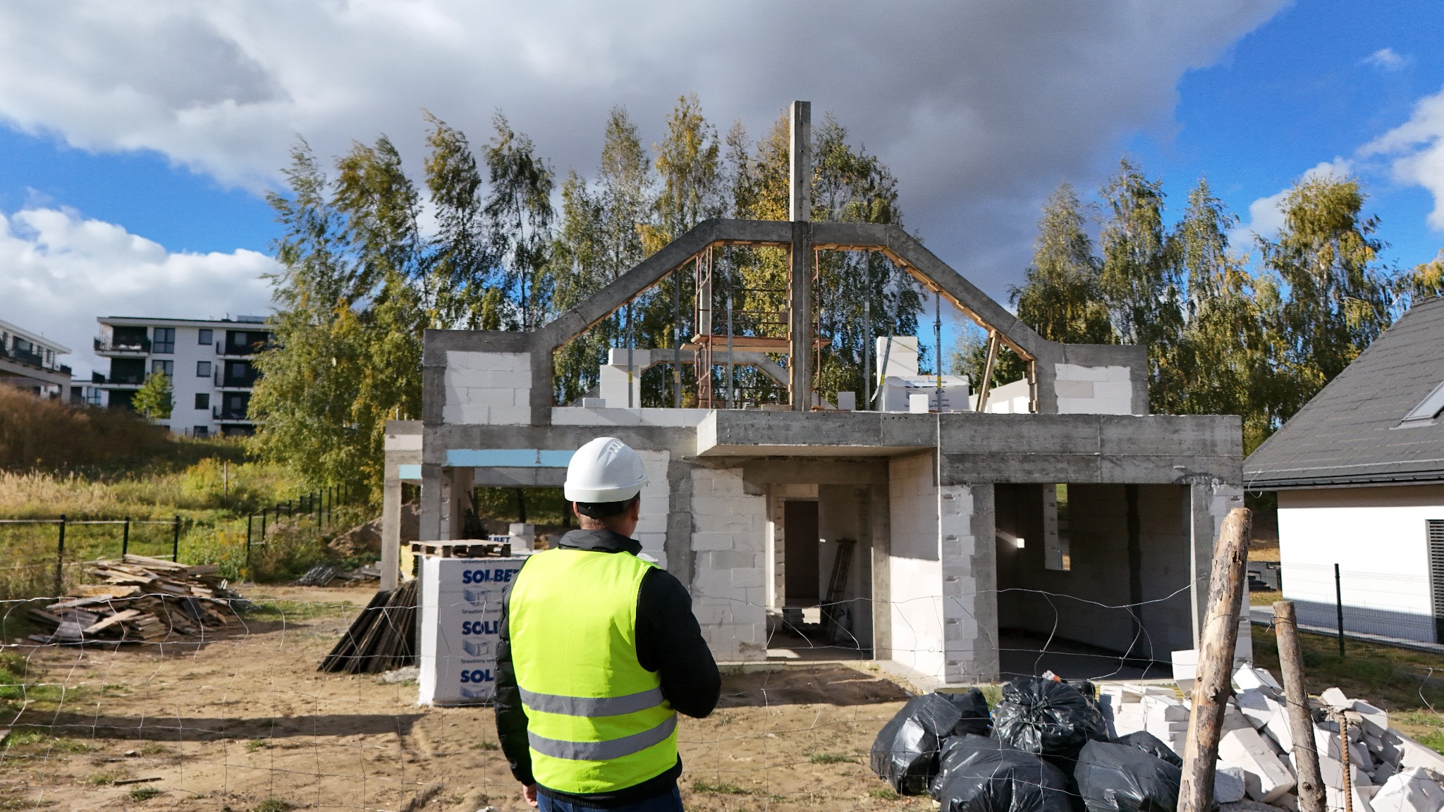 Budowa domu jednorodzinnego w stanie surowym otwartym. Widoczny pracownik w kamizelce odblaskowej i kasku. W tle drzewa i błękitne niebo z chmurami.