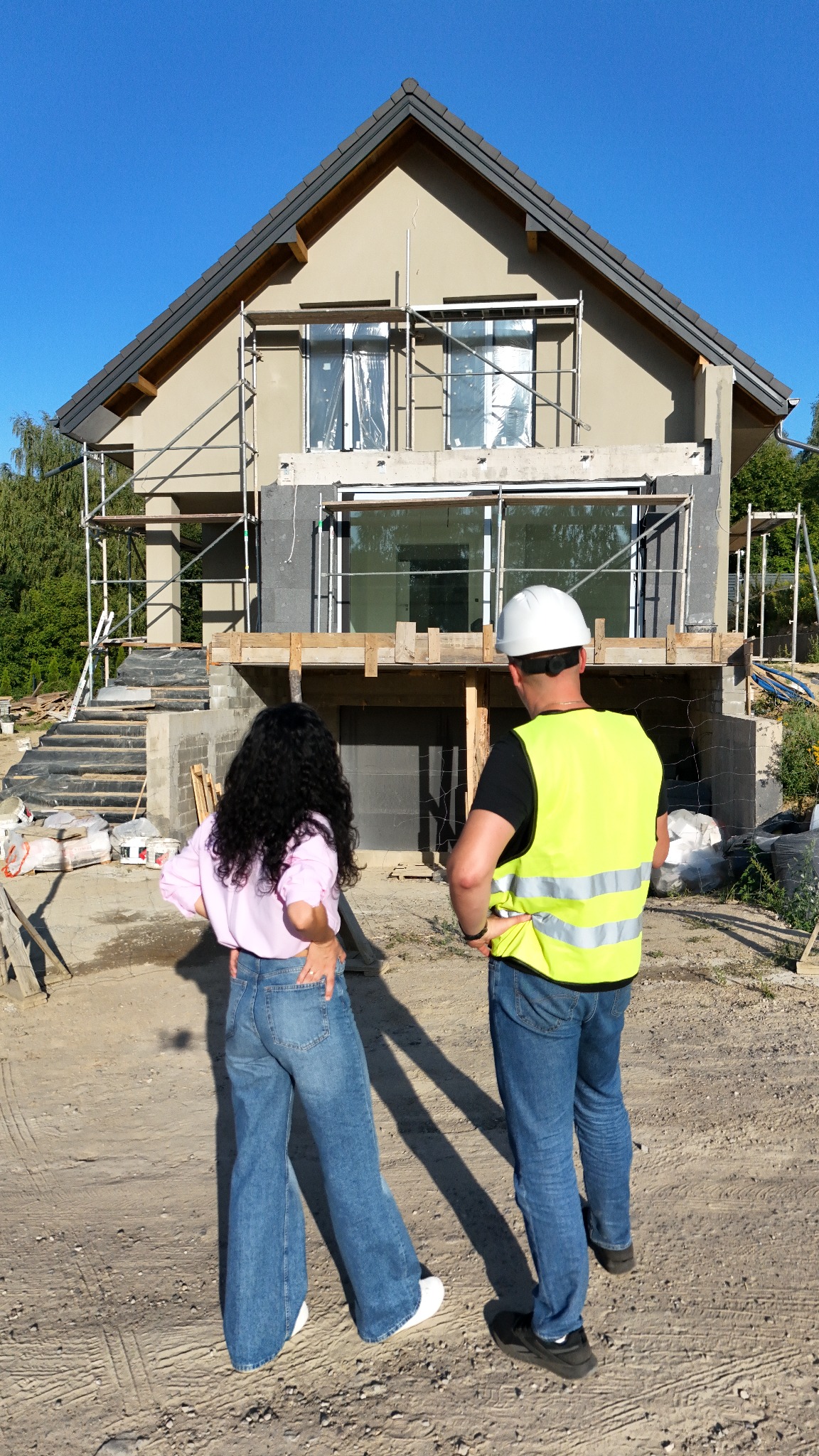 Dwie osoby obserwują budowę domu jednorodzinnego z rusztowaniami. Widoczne okna zabezpieczone folią i surowy stan elewacji. Osoba w kamizelce odblaskowej i kasku.