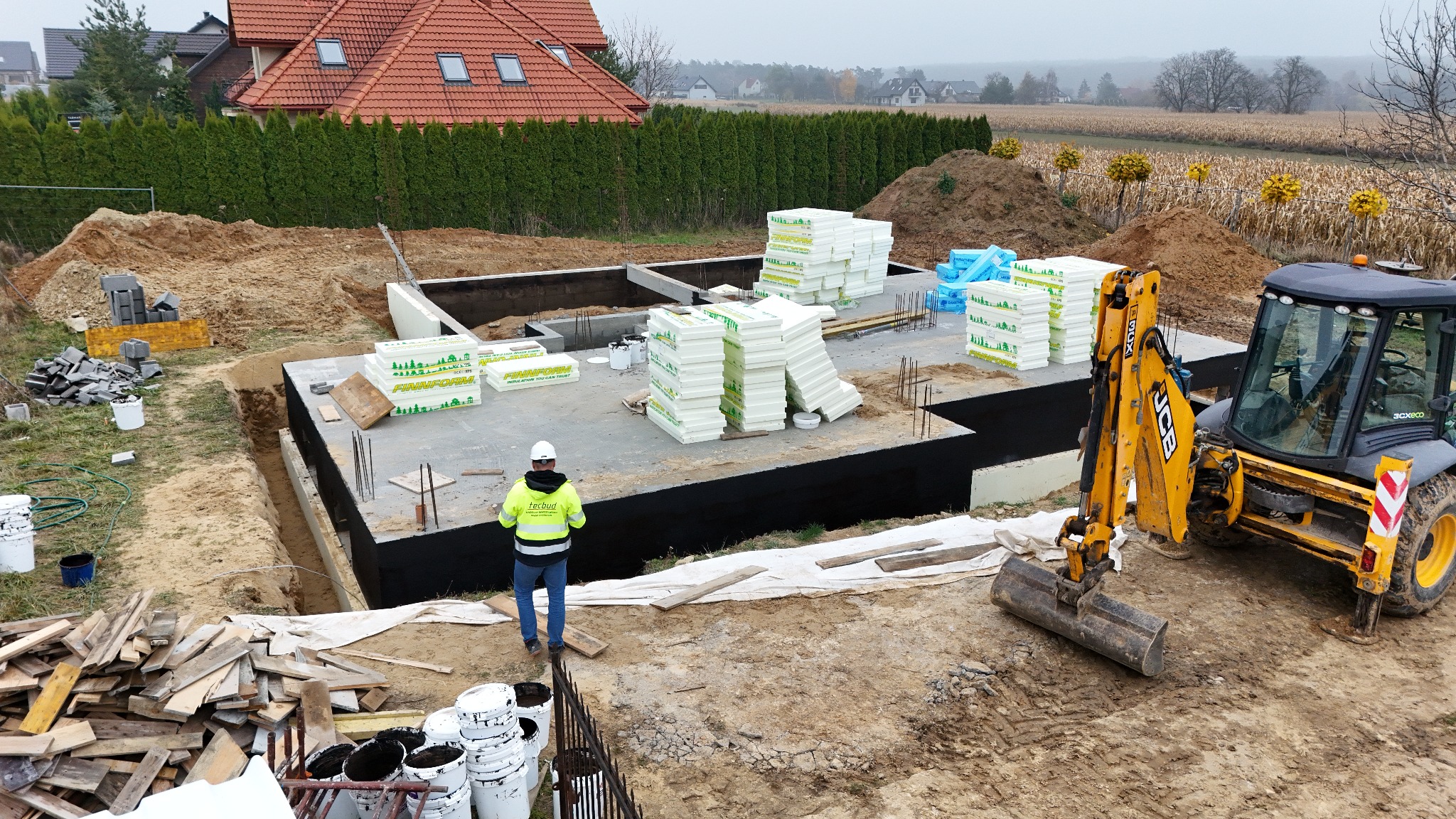 Fundamenty domu jednorodzinnego w trakcie budowy, zabezpieczone izolacją, z widocznym pracownikiem w kasku i koparką JCB. W tle dom i pole uprawne.