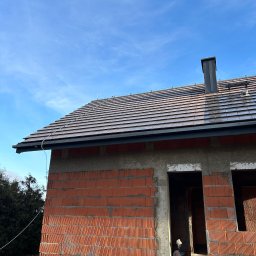 PRZEDSIEBIORSTWO TRANSPORTOWO - BUDOWLANE '' SIGMA'' RYSZARD TRZYNOGA - Surowy dom z czerwonej cegły z ukończonym dachem z ciemnej dachówki i czarną rynną, na tle błękitnego nieba. Widoczne detale murarskie i komin.