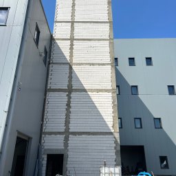 PRZEDSIEBIORSTWO TRANSPORTOWO - BUDOWLANE '' SIGMA'' RYSZARD TRZYNOGA - Wysoka ściana z białych bloczków, z widocznymi spoinami i zbrojeniami, w trakcie budowy. Obok budynek z szarej blachy trapezowej i niebieskie niebo w tle.