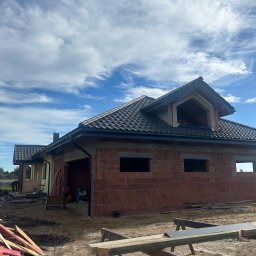 PRZEDSIEBIORSTWO TRANSPORTOWO - BUDOWLANE '' SIGMA'' RYSZARD TRZYNOGA - Dobudowa domu z cegły z ciemnym dachem pod błękitnym niebem. Widoczne rusztowania i materiały budowlane. Częściowo otynkowana elewacja.