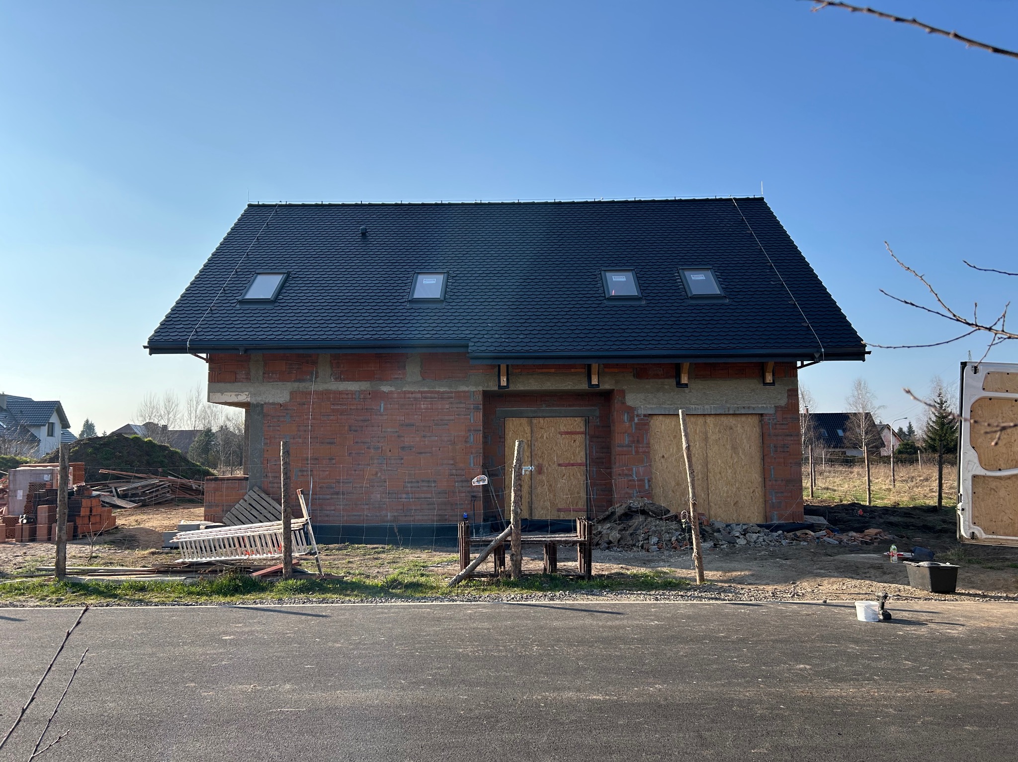 Niedokończony dom z czerwonej cegły z czarnym dachem i trzema oknami dachowymi, zabezpieczone wejście deskami, widok z ulicy w słoneczny dzień.