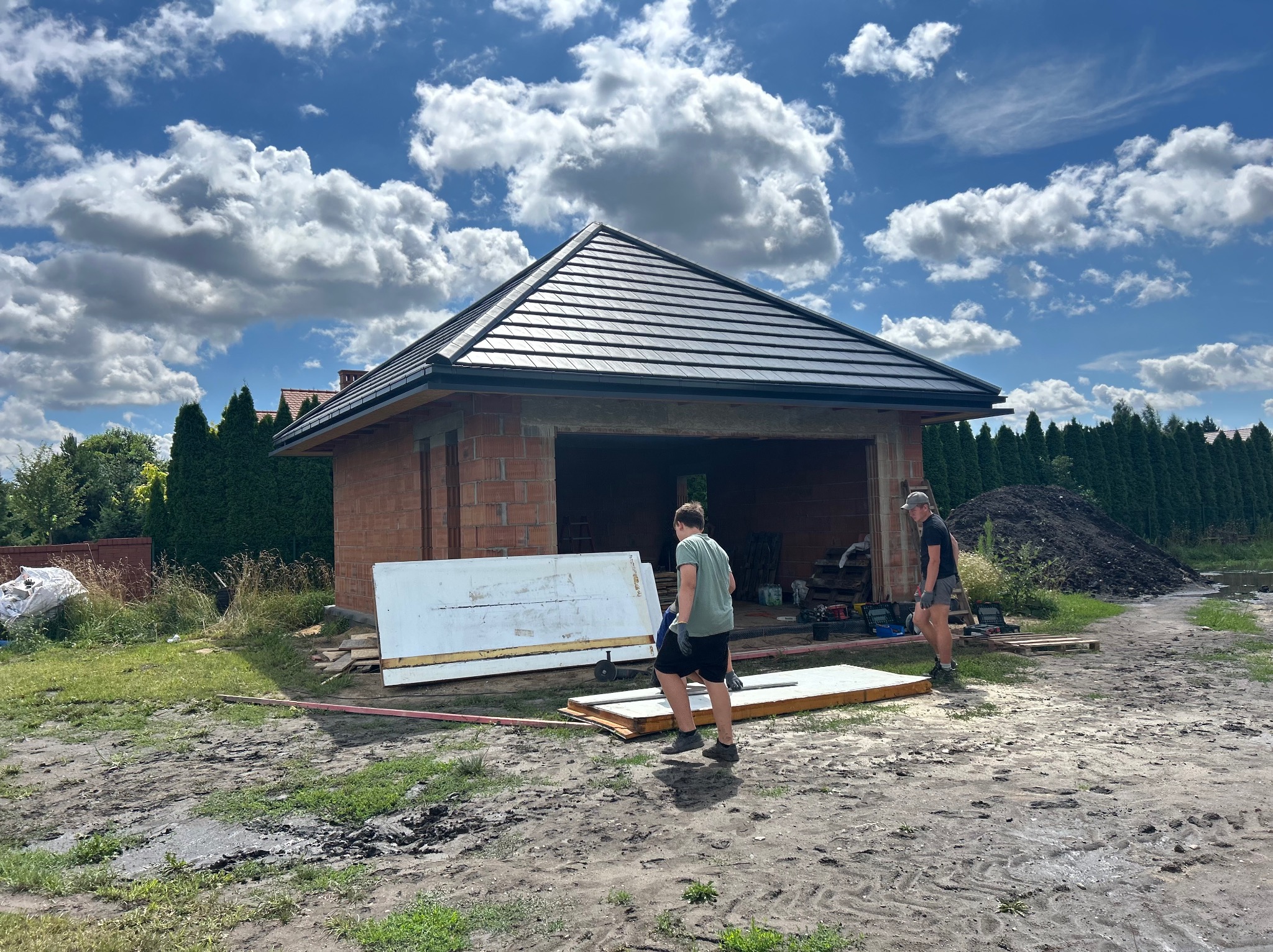 Budowa domu jednorodzinnego z garażem w trakcie realizacji. Widoczny budynek z czerwonej cegły z czarnym dachem, pracownicy przenoszą płyty. Błękitne niebo z chmurami.