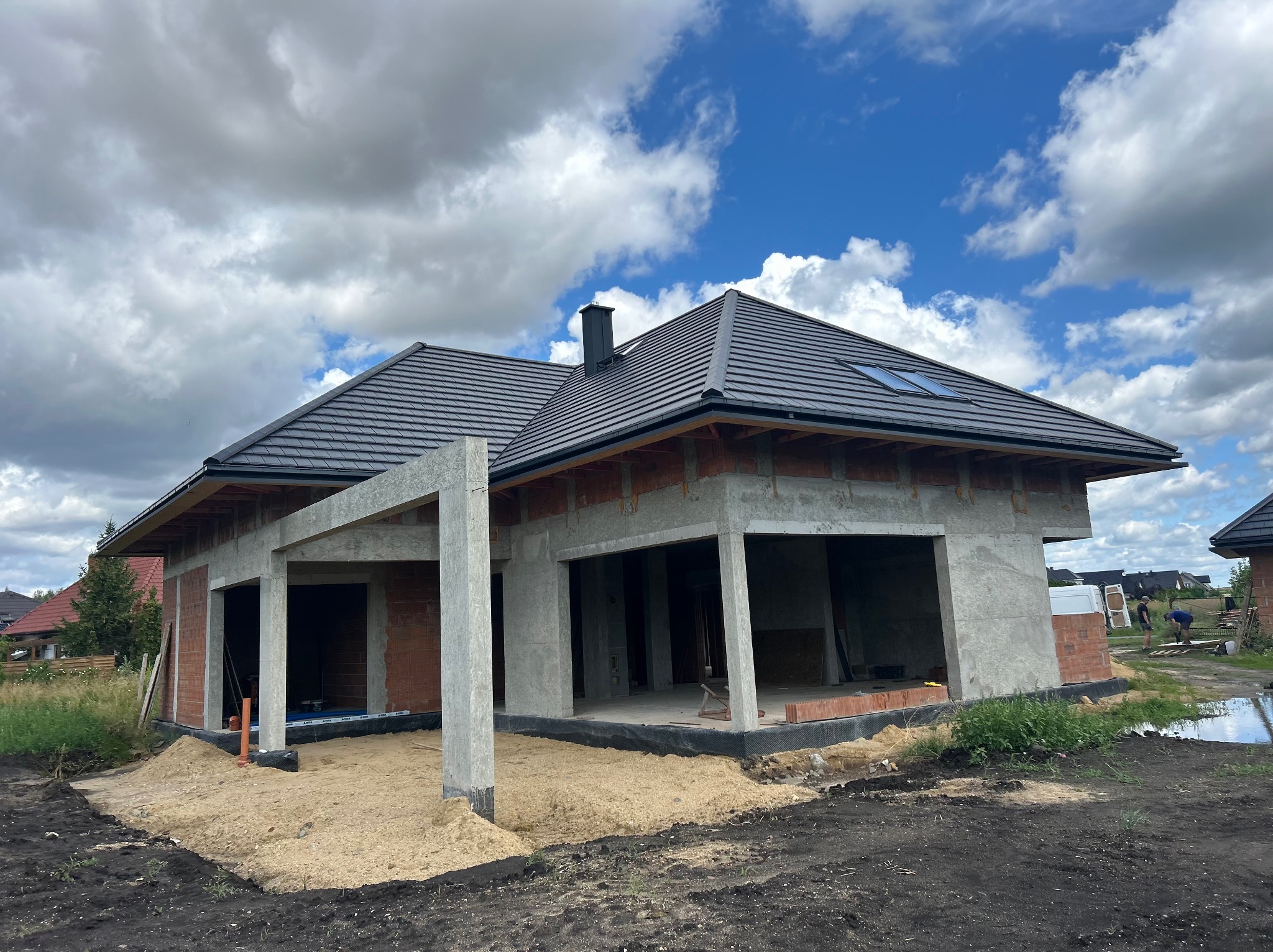 Dom jednorodzinny w trakcie budowy z garażem, widoczne ściany z betonu komórkowego, dach z dachówki ceramicznej i fragment nieba z chmurami.