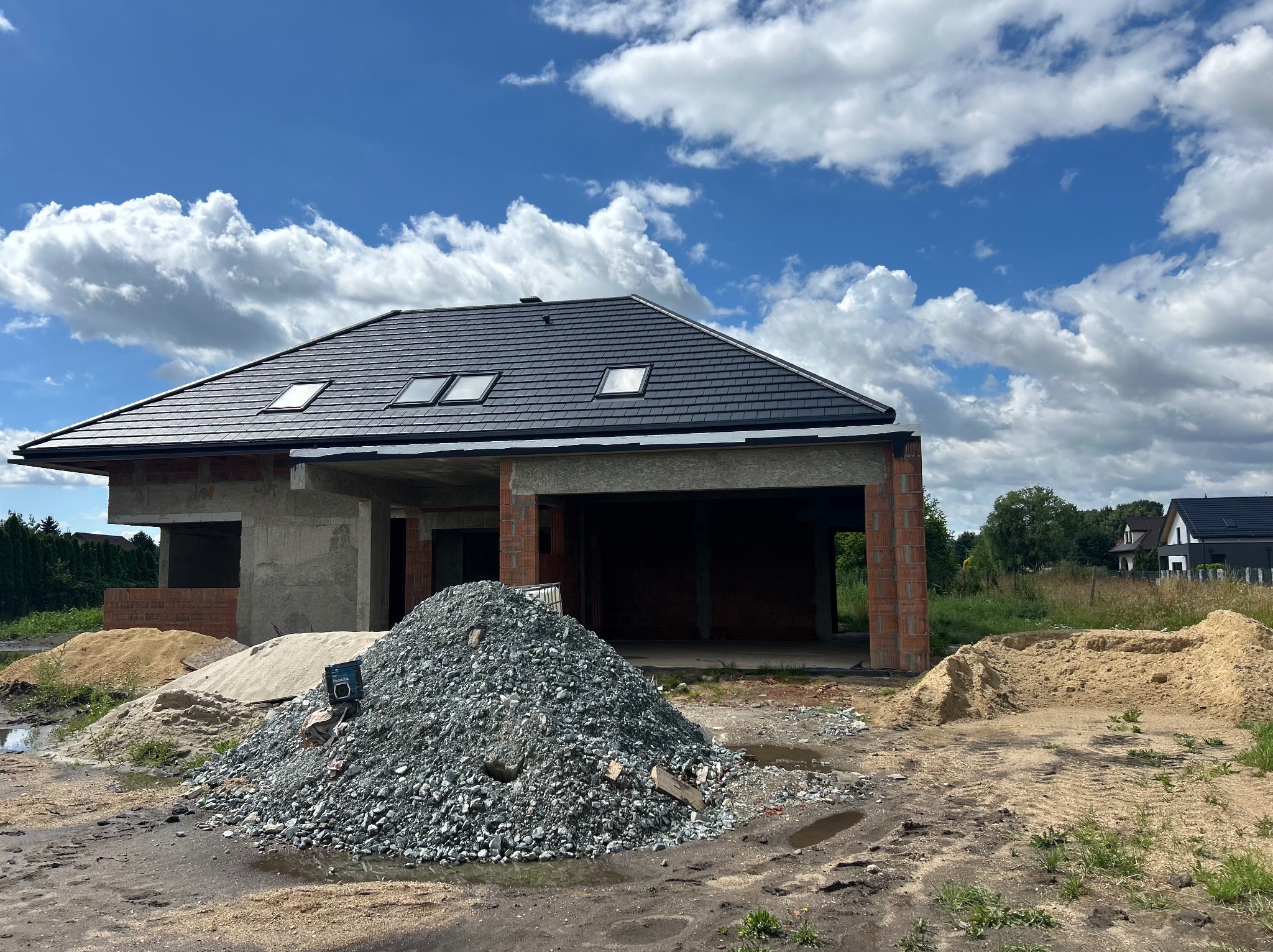 Dom jednorodzinny w trakcie budowy z czarnym dachem i trzema oknami dachowymi. Na pierwszym planie hałda gruzu i piasku. Błękitne niebo z białymi chmurami w tle.