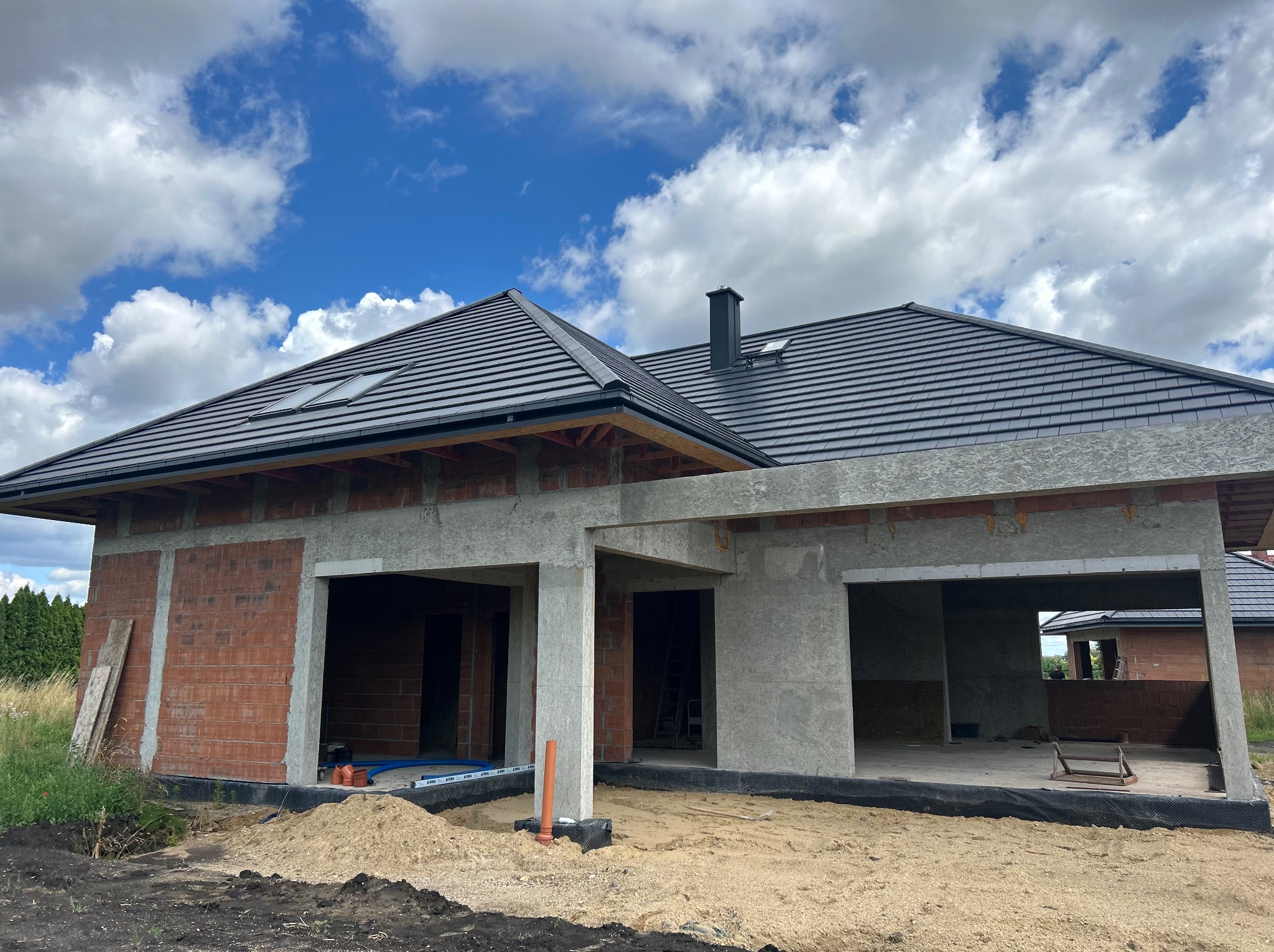 Dom jednorodzinny w trakcie budowy: murowane ściany z pustaków, czarny dach z dachówką, widoczne elementy więźby, błękitne niebo z chmurami w tle. Stan surowy otwarty.