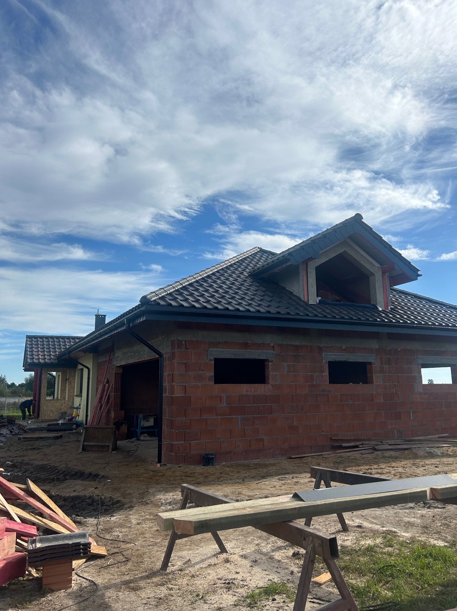 Dobudowa domu z cegły z ciemnym dachem pod błękitnym niebem. Widoczne rusztowania i materiały budowlane. Częściowo otynkowana elewacja.