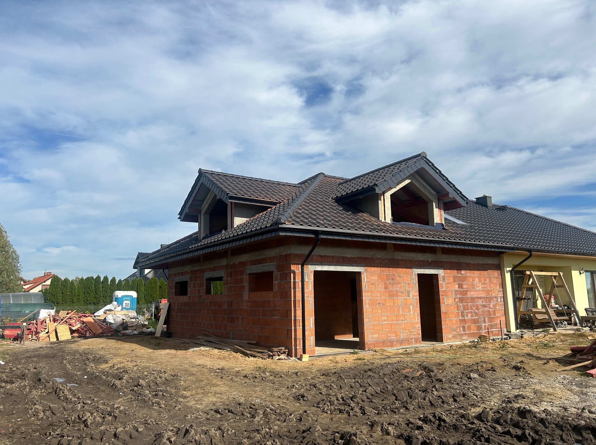 Budowa domu z czerwonej cegły, widoczne otwory okienne i drzwiowe, dach pokryty ciemną dachówką. Na pierwszym planie ziemia, w tle błękitne niebo z chmurami.