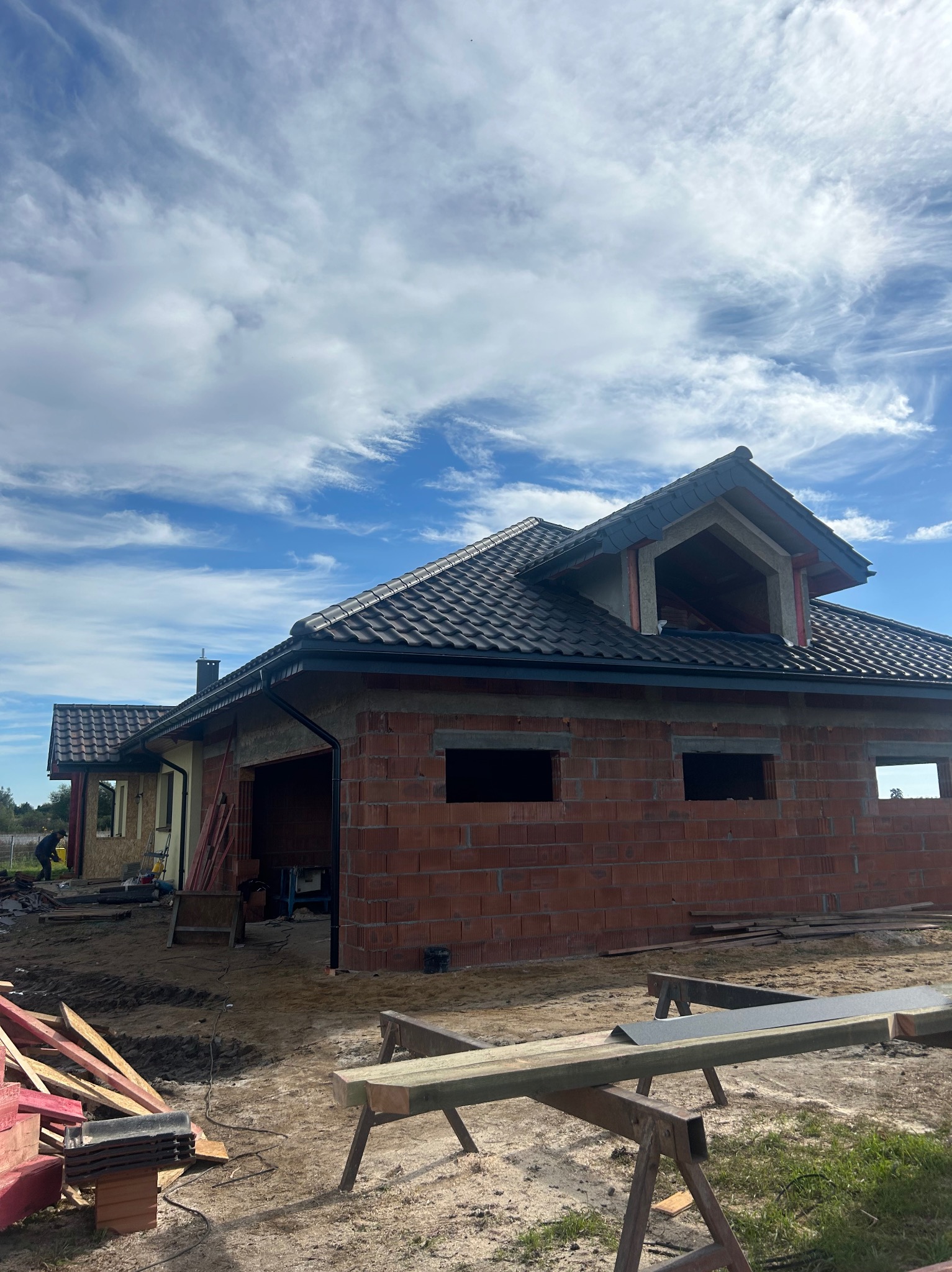 Budowa domu jednorodzinnego z czerwonej cegły, z widocznym dachem pokrytym ciemną dachówką. W tle błękitne niebo z chmurami. Widoczne rusztowania i materiały budowlane.