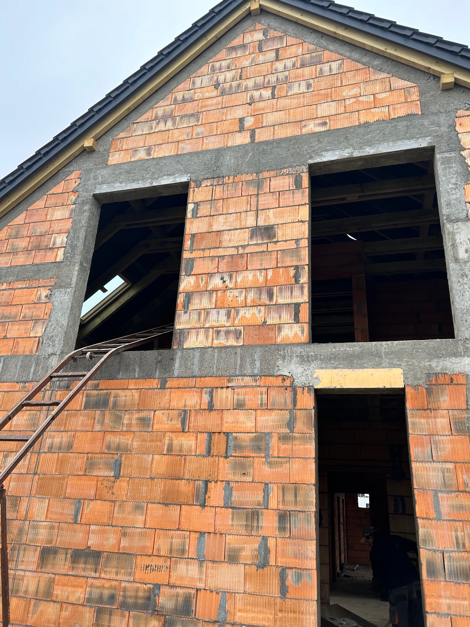 Surowy stan domu z czerwonej cegły z otworami okiennymi i drabiną. Widoczna konstrukcja dachu i fragment wnętrza. Etap budowy domu jednorodzinnego.