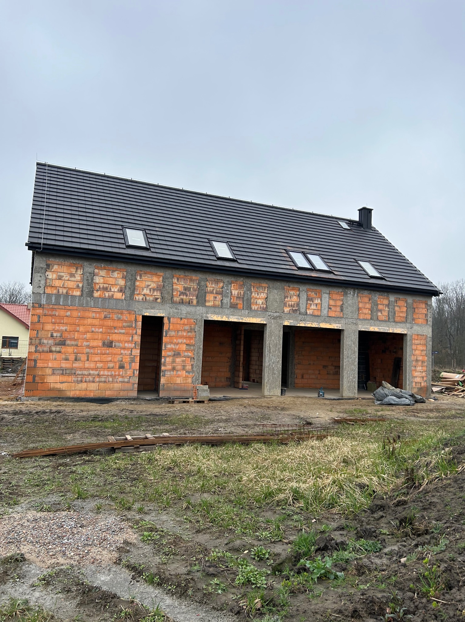 Surowy stan domu z czerwonej cegły z ciemnym dachem i oknami dachowymi. Widoczne otwory okienne i drzwiowe. Teren wokół domu częściowo błotnisty.