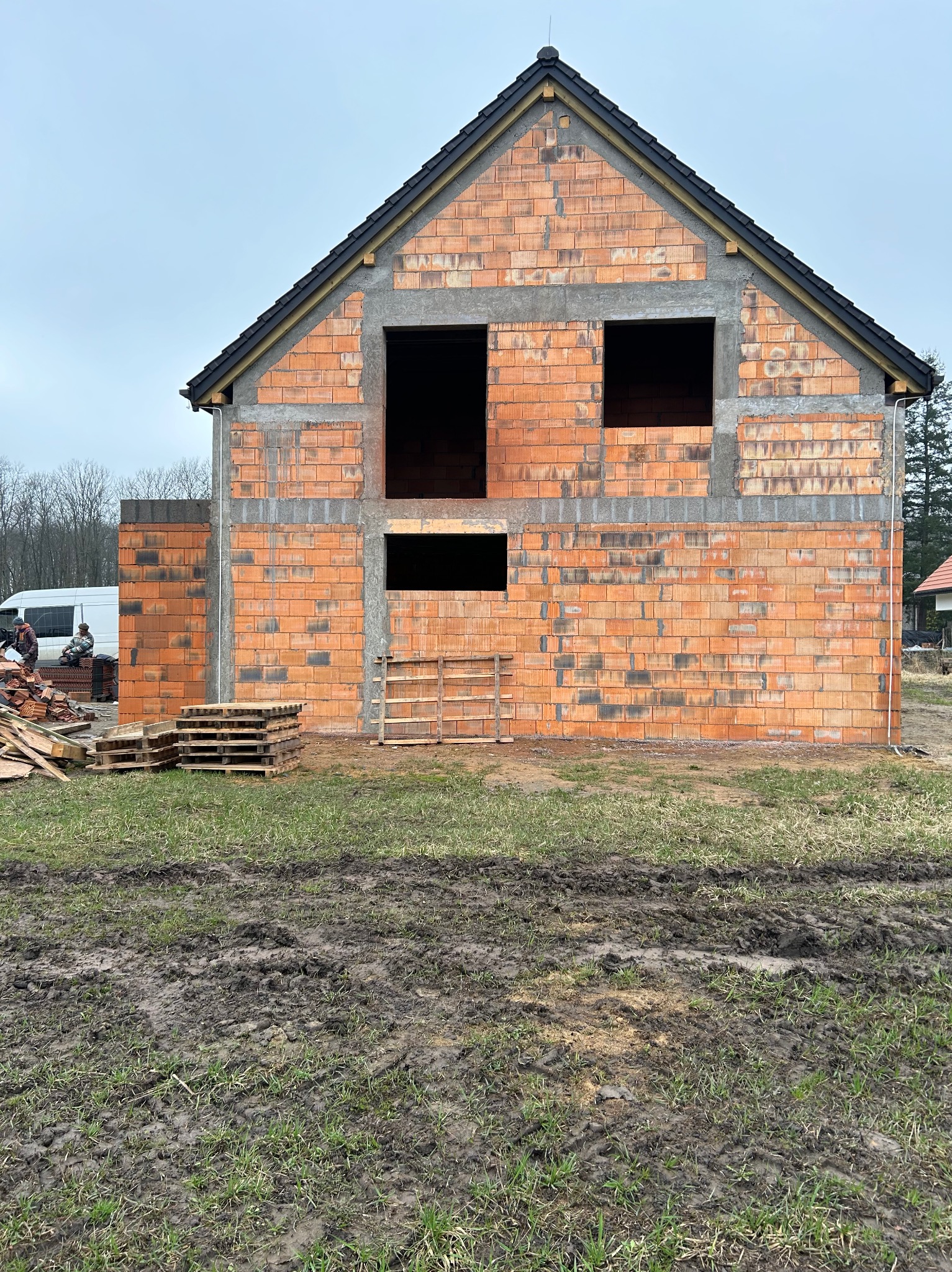 Surowy stan domu z czerwonej cegły z widocznymi otworami okiennymi i zarysem dachu. Na pierwszym planie palety i błotniste podłoże. W tle częściowo widoczny bus.