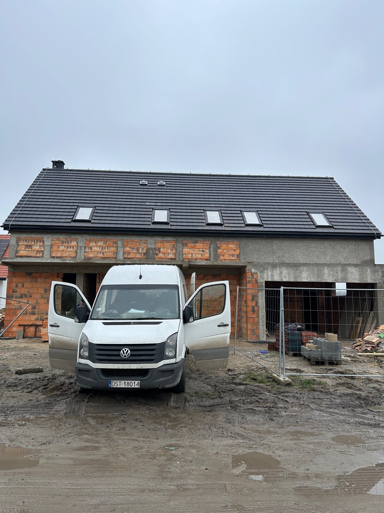 Dom w trakcie budowy z częściowo położonym dachem i oknami dachowymi, na pierwszym planie biały bus z otwartymi drzwiami na błotnistym terenie.