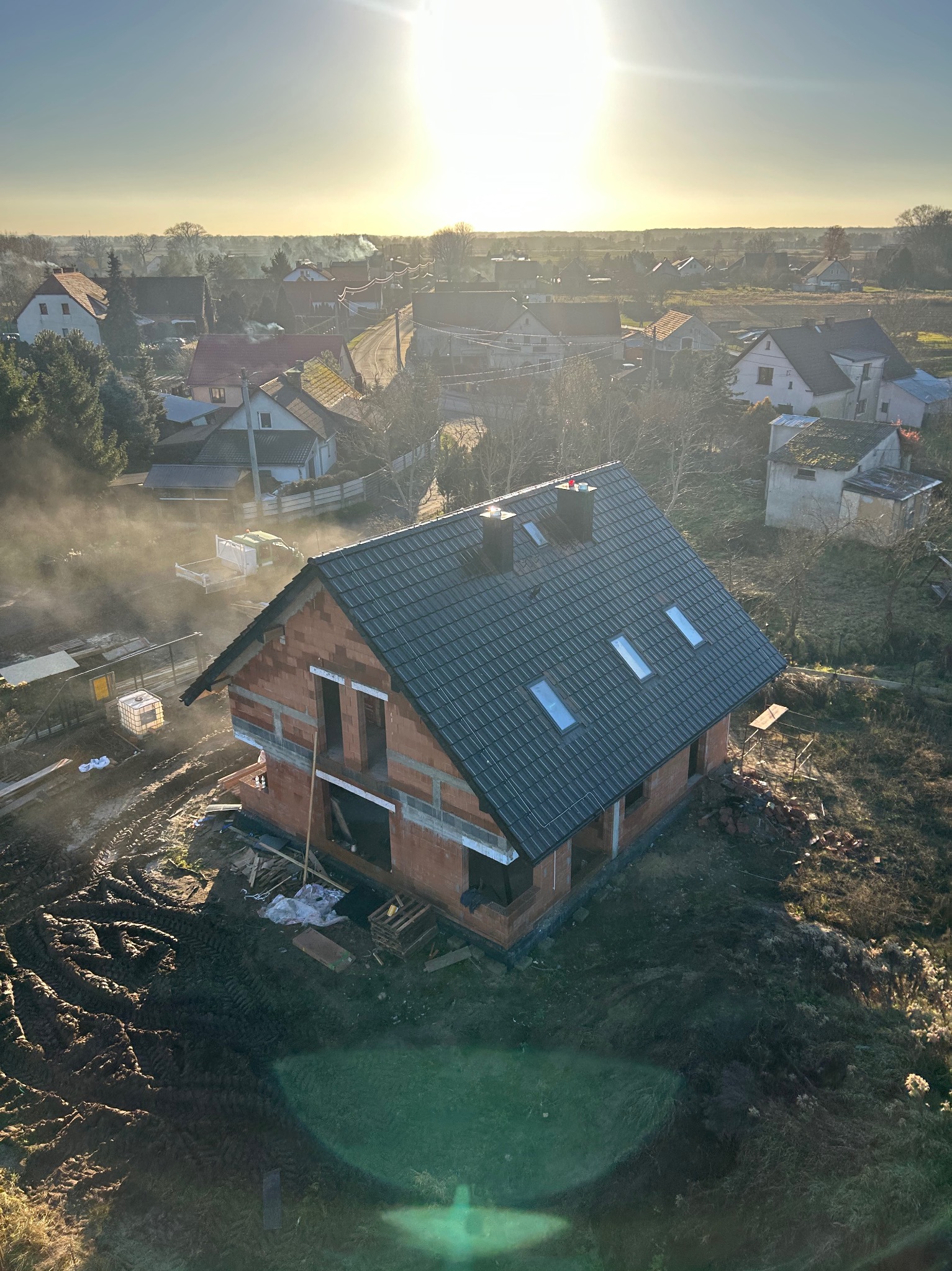 Wysoki kąt na dom w trakcie budowy z ciemną dachówką i ceglanymi ścianami, otoczony błotnistym terenem i innymi domami w tle, w słoneczny dzień.