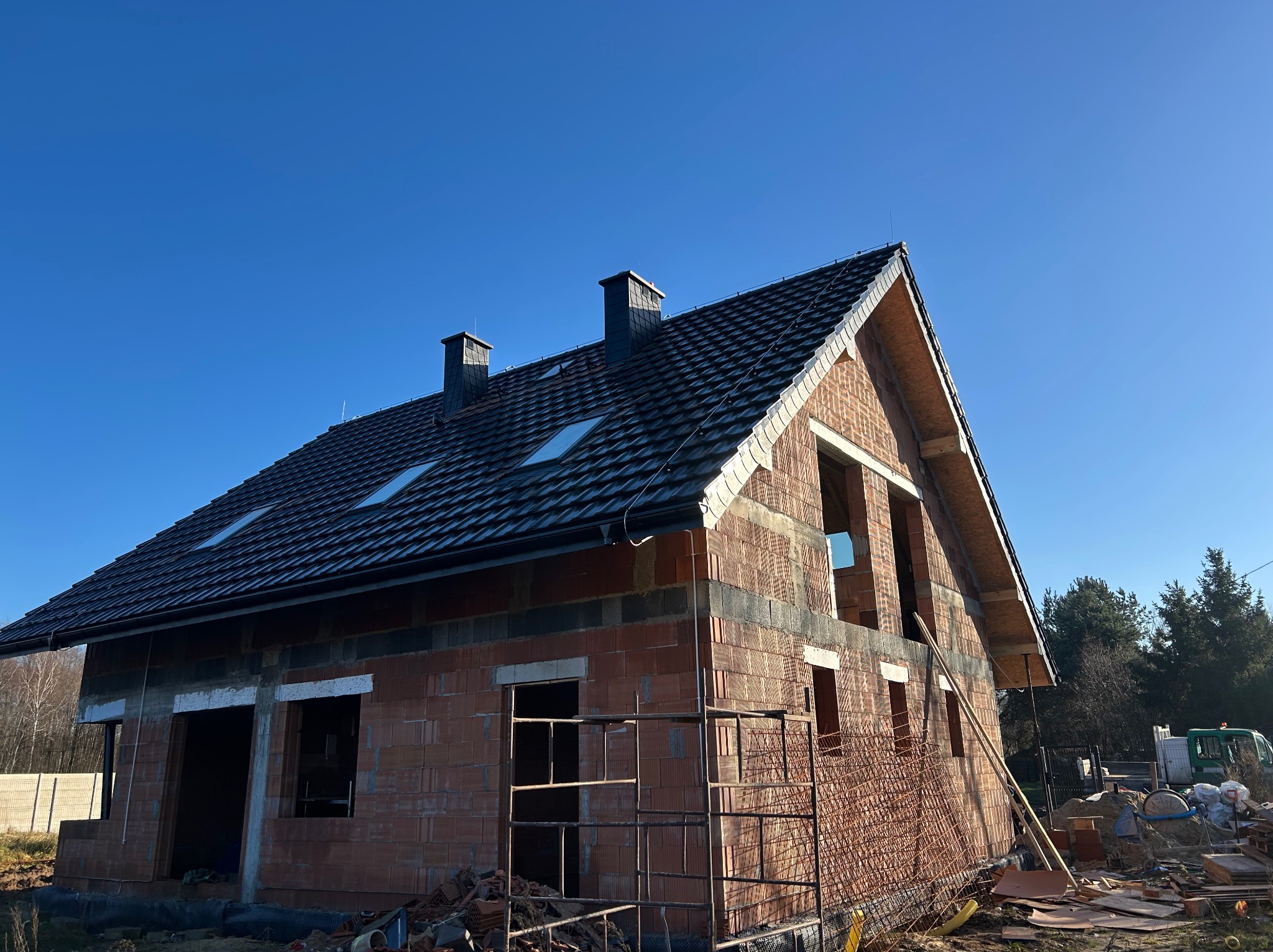 Dom w trakcie budowy z ciemnym dachem i ceglanymi ścianami. Widoczne okna dachowe i kominy. Na pierwszym planie rusztowanie i materiały budowlane.