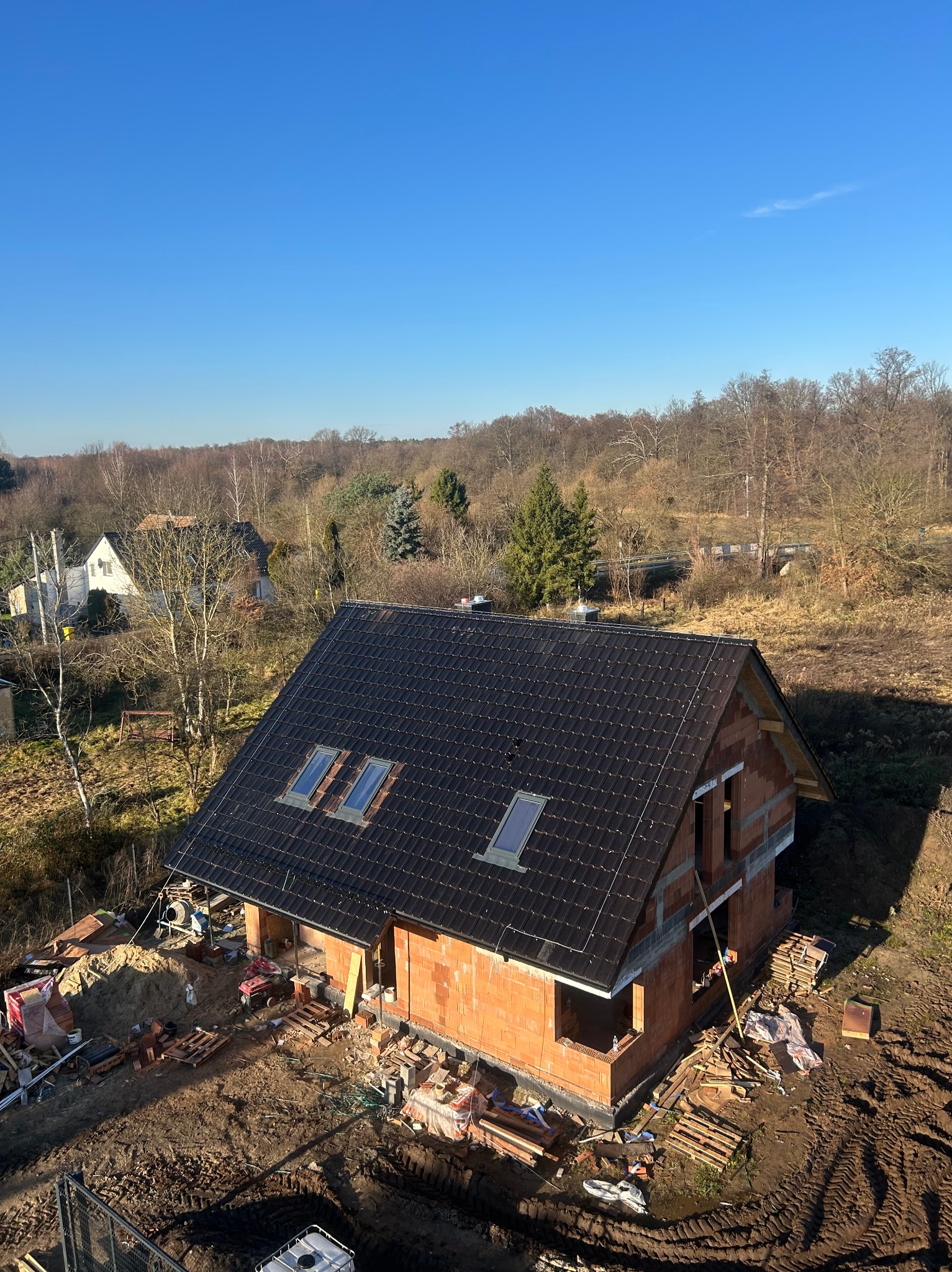Dom w trakcie budowy z ciemnym dachem i oknami dachowymi, murowane ściany z czerwonej cegły, otoczenie placu budowy, widok z góry w słoneczny dzień.