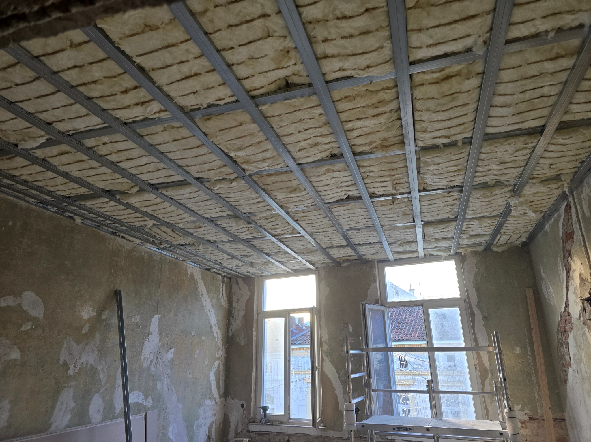 Montaż izolacji z wełny mineralnej na metalowym ruszcie sufitowym w remontowanym pokoju z oknami i rusztowaniem. Widoczne ślady po starych tynkach.