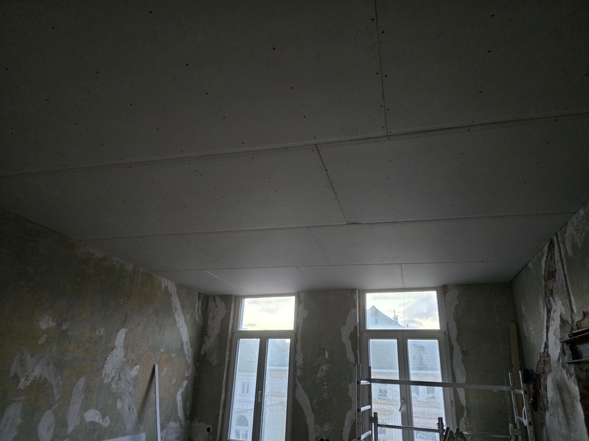 Pokój w trakcie remontu: sufit z płyt karton-gips, ściany częściowo przygotowane do malowania, widoczne okna i rusztowanie. Prace wykończeniowe w starym budownictwie.