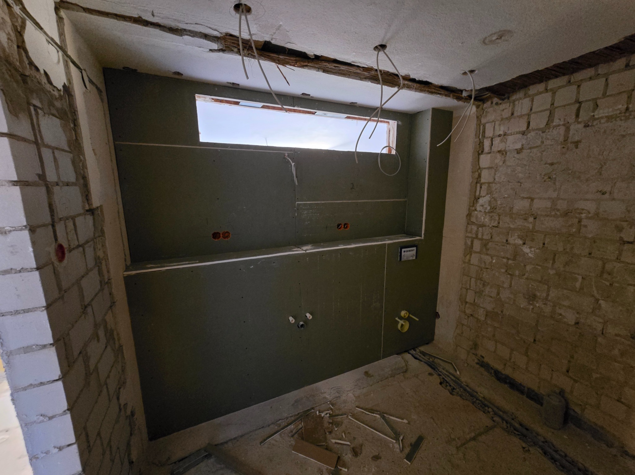 Ściana z płyty karton-gips z oknem w trakcie montażu, widoczne instalacje i surowa cegła. Prace wykończeniowe w łazience, widok na otwór okienny.