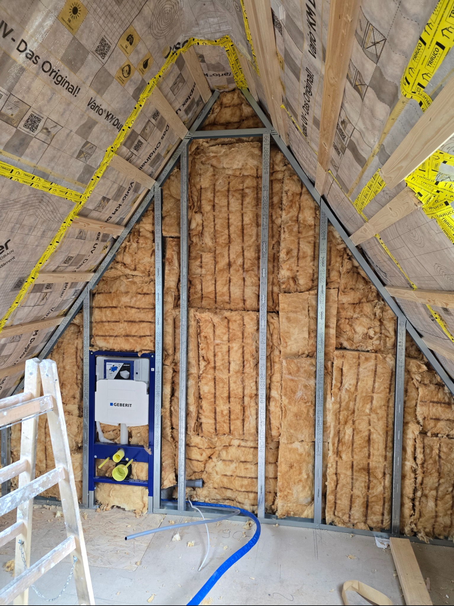 Skośna ściana poddasza w trakcie izolacji wełną mineralną, widoczne profile metalowe, folia paroizolacyjna i stelaż pod zabudowę WC Geberit. Prace wykończeniowe.