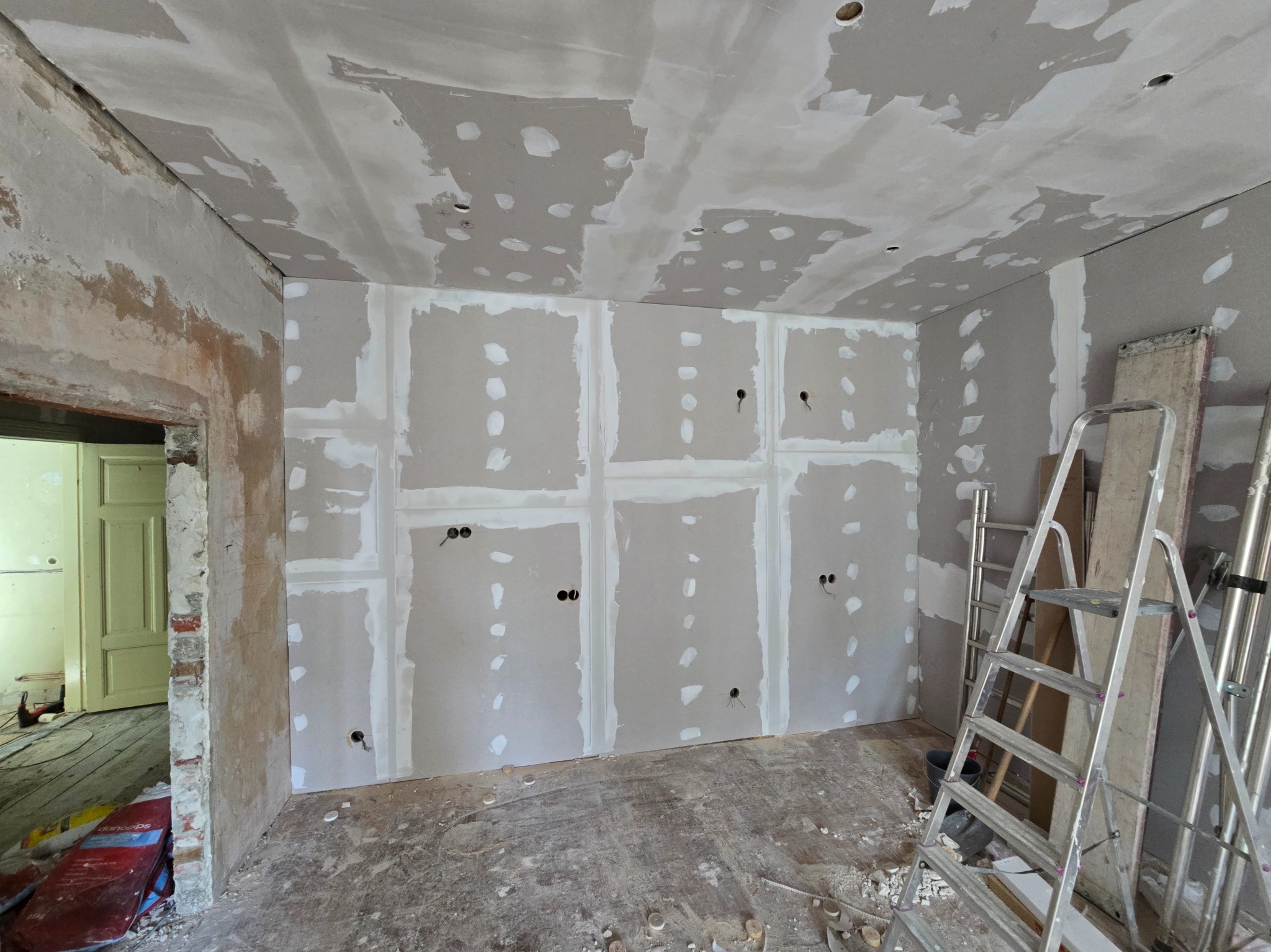 Pokój w trakcie prac: ściany obłożone płytami gipsowo-kartonowymi, widoczne otwory na gniazdka, częściowo wyszpachlowane łączenia, drabina oparta o ścianę.