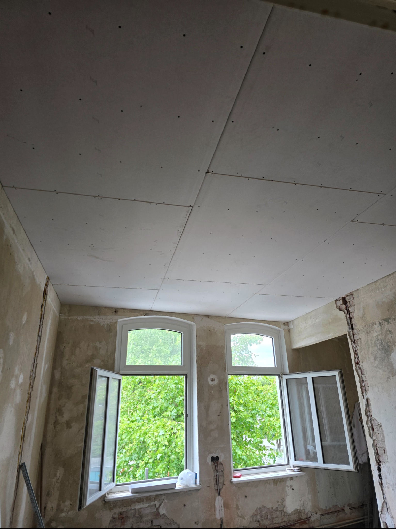 Montaż płyt karton-gips na suficie w remontowanym pokoju. Widoczne otwarte okna z zielenią na zewnątrz i surowe ściany w trakcie prac wykończeniowych.