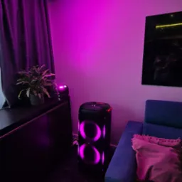 Pokój oświetlony fioletowym światłem z głośnika JBL Partybox, stojącego obok niebieskiej sofy z poduszką. Na szafce roślina doniczkowa i mały reflektor. Nowoczesne oświetlenie.