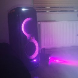 ZestawPartyKrk - Głośnik JBL Partybox z pulsującym, fioletowym oświetleniem LED, obok mały reflektor, tworzący nastrojową atmosferę w pomieszczeniu z grzejnikiem i rośliną w tle.