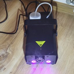 ZestawPartyKrk - Czarny wytwornica dymu z fioletowym światłem na drewnianej podłodze, widoczny przewód zasilający i zbiornik na płyn. Urządzenie oświetleniowe na imprezę.