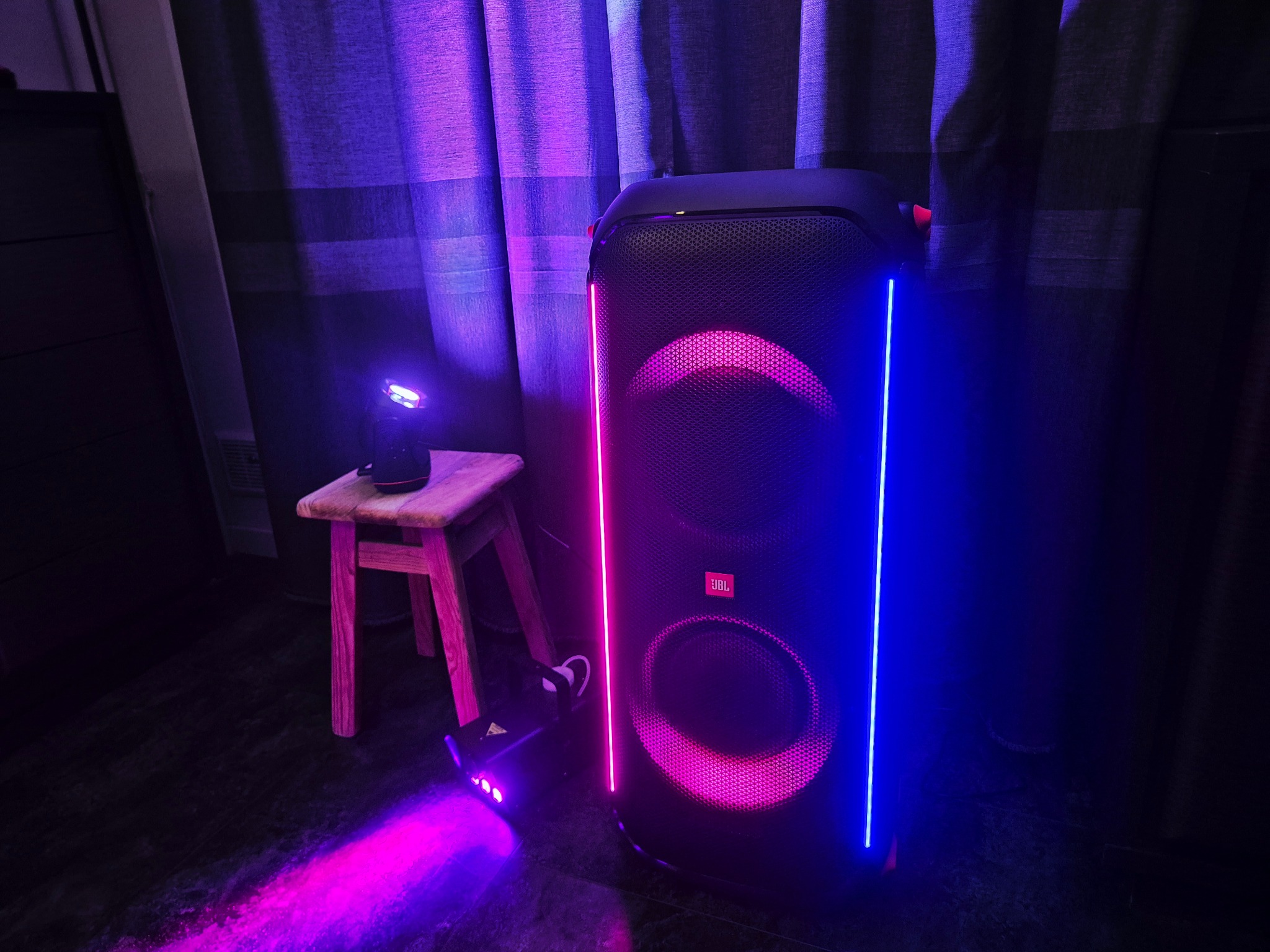 Głośnik JBL PartyBox z podświetleniem LED w odcieniach różu i niebieskiego, ustawiony obok małego stołka z reflektorem, tworzący klubową atmosferę w zaciemnionym pomieszczeniu.