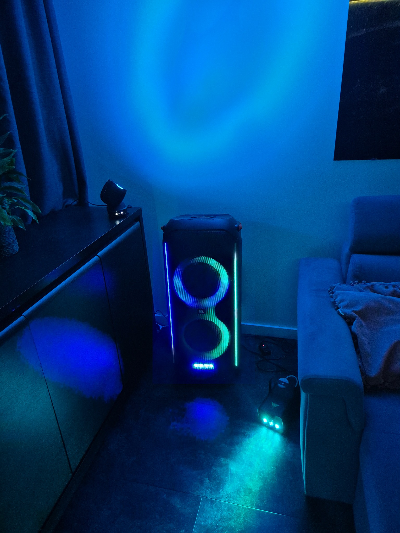 Kolumna głośnikowa JBL Partybox z podświetleniem LED w odcieniach niebieskiego i zielonego, ustawiona w rogu pokoju obok sofy i szafki. Dodatkowo widoczna wytwornica dymu.