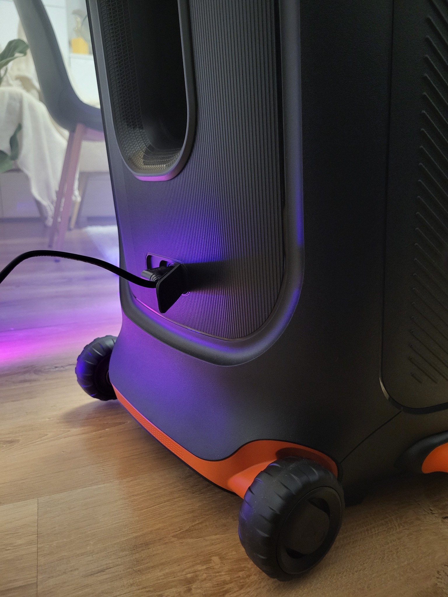 Mobilny głośnik imprezowy z podświetleniem LED, czarno-pomarańczowa obudowa na kółkach, kabel zasilający podłączony, fioletowa poświata na podłodze.
