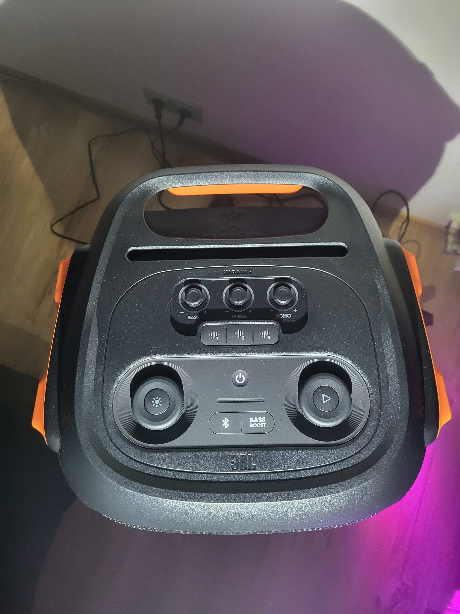 Czarno-pomarańczowy głośnik JBL Partybox z widocznymi przyciskami i podświetleniem, ustawiony na podłodze, gotowy do użycia podczas imprezy.