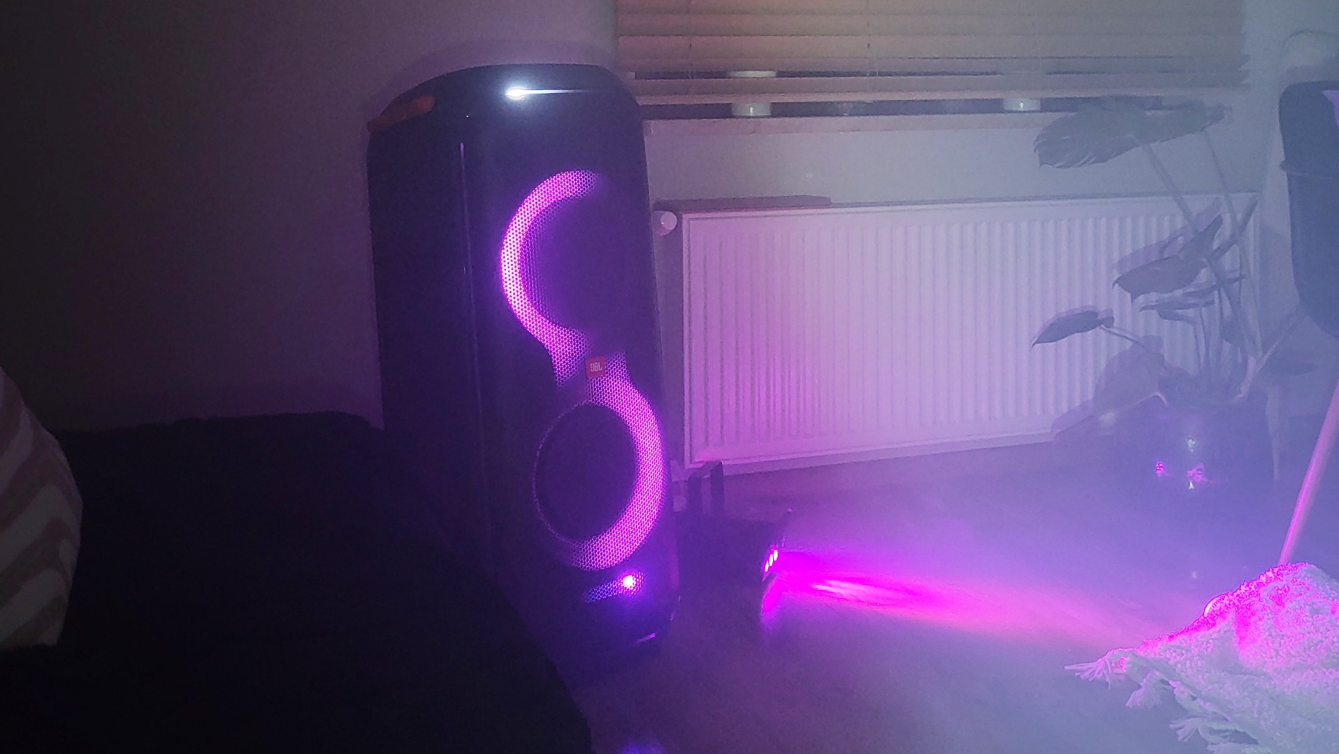 Głośnik JBL Partybox z pulsującym, fioletowym oświetleniem LED, obok mały reflektor, tworzący nastrojową atmosferę w pomieszczeniu z grzejnikiem i rośliną w tle.