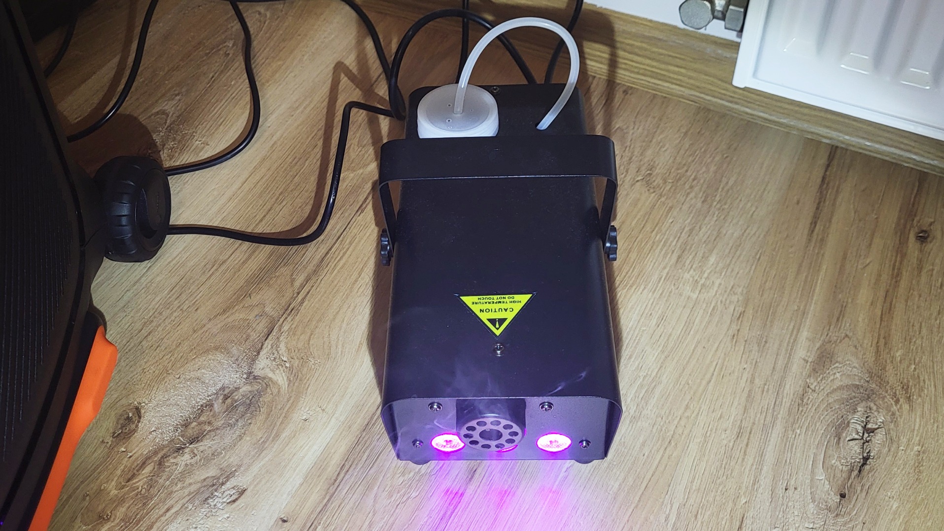 Czarny wytwornica dymu z fioletowym światłem na drewnianej podłodze, widoczny przewód zasilający i zbiornik na płyn. Urządzenie oświetleniowe na imprezę.