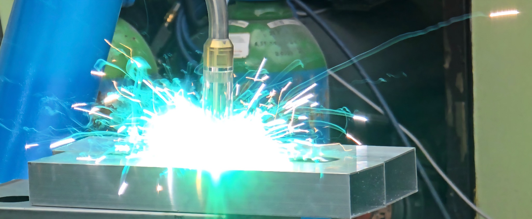 Intensywne iskry podczas spawania grubego, srebrnego bloku metalu. Widoczny palnik spawalniczy i butle z gazem w tle. Jasne, niebiesko-białe światło spawania.