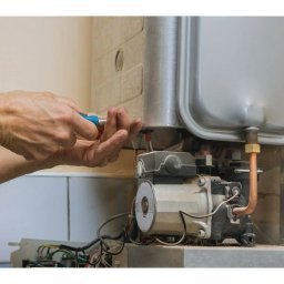 INSTALATOR S.C. KAZIMIERZ OZIMEK WOJCIECH OZIMEK - Serwisant naprawia piec gazowy, używając śrubokręta. Widoczne elementy pieca, rury miedziane i dłonie montera podczas pracy. Ujęcie z bliska, detale instalacji.