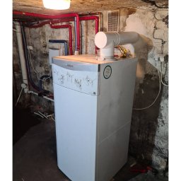 INSTALATOR S.C. KAZIMIERZ OZIMEK WOJCIECH OZIMEK - Biały piec gazowy Vaillant w piwnicy z widocznymi rurami instalacji grzewczej i oświetleniem sufitowym. Ściany piwnicy z surowym wykończeniem.