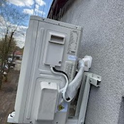 INSTALATOR S.C. KAZIMIERZ OZIMEK WOJCIECH OZIMEK - Boczny widok zamontowanej jednostki zewnętrznej klimatyzacji na elewacji budynku z kolcami przeciw ptakom na dachu. Widoczne przewody i uchwyty montażowe.