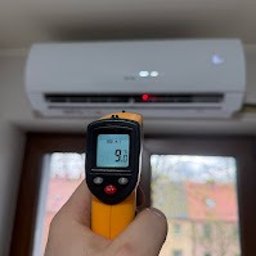 INSTALATOR S.C. KAZIMIERZ OZIMEK WOJCIECH OZIMEK - Pomiar temperatury nawiewu klimatyzatora termometrem elektronicznym. Widoczny biały klimatyzator na ścianie i okno w tle. Precyzyjny pomiar chłodzenia.