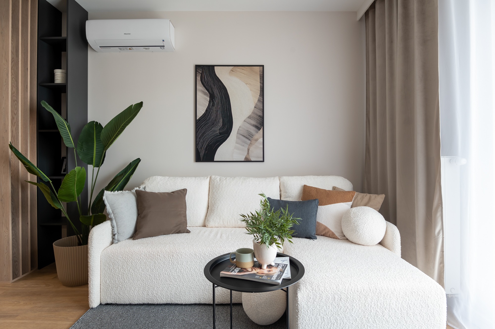 Jasne wnętrze salonu z sofą boucle, obrazem abstrakcyjnym i roślinami. Widoczny klimatyzator i zasłony w neutralnych barwach. Minimalistyczny design z akcentami natury.