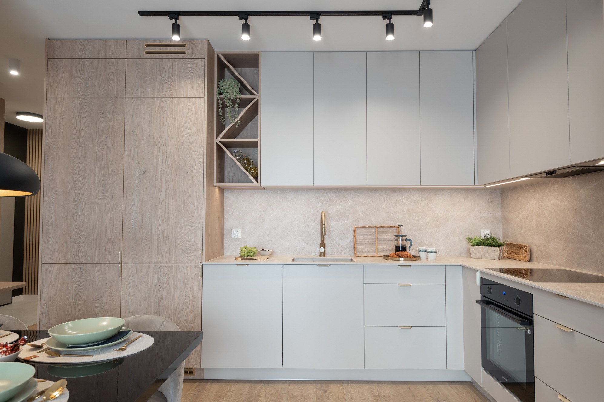 Jasna, nowoczesna kuchnia z drewnianymi i szarymi szafkami, złotą armaturą i oświetleniem punktowym. Na blacie dekoracje i naczynia, stół z zastawą w tle.