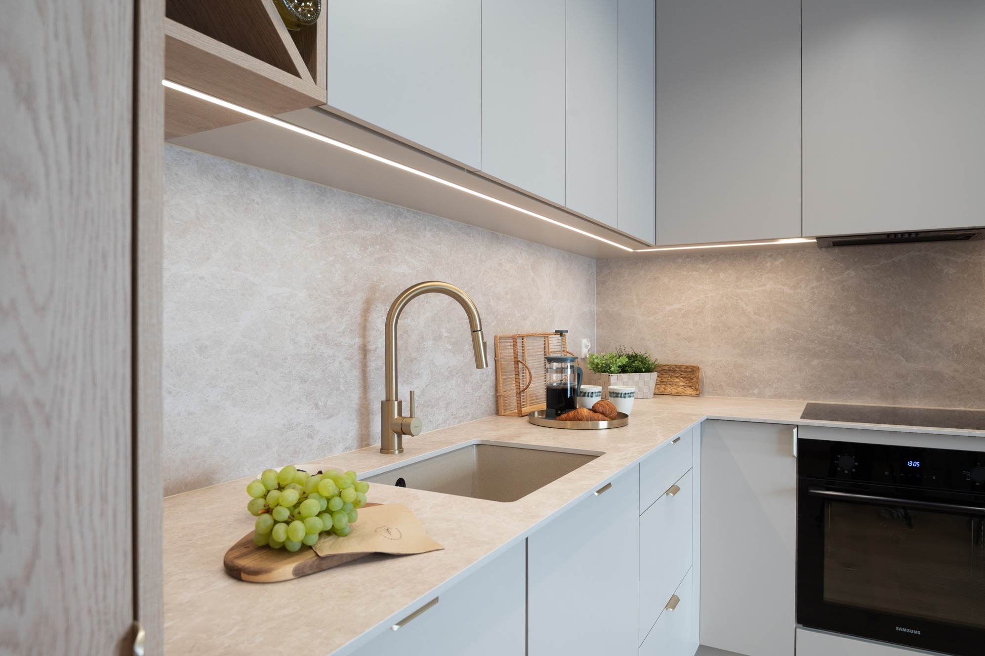 Jasna kuchnia w stylu minimalistycznym z blatem imitującym marmur, złotą armaturą i oświetleniem LED pod szafkami. Na blacie winogrona i śniadanie.