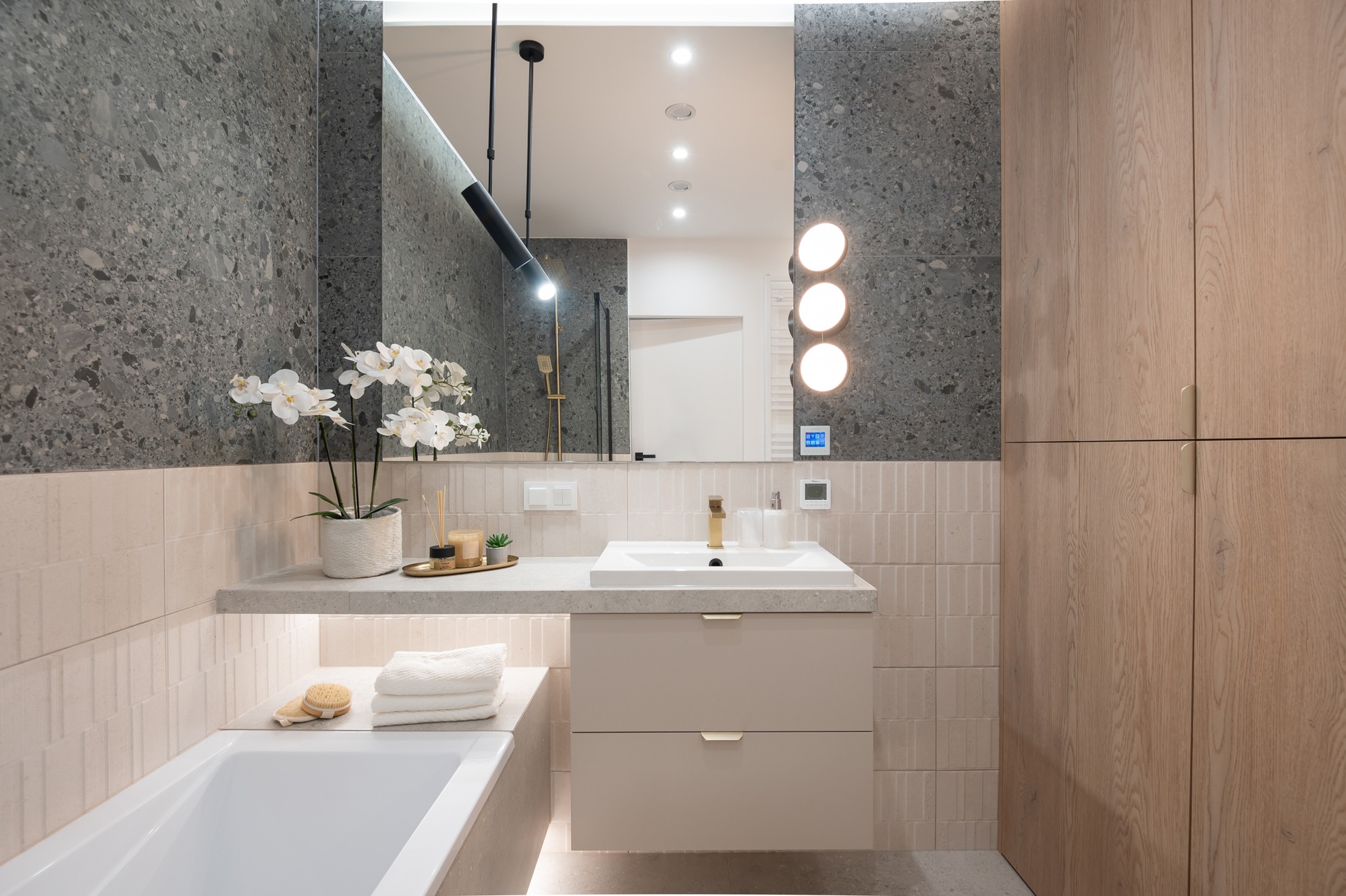 Nowoczesna łazienka z szarą mozaiką, beżowymi płytkami i drewnianymi szafkami. Umywalka z szafką w kolorze beżowym, lustro z oświetleniem i dekoracje.